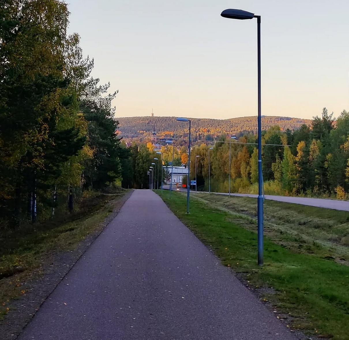 En asfalterad gång och cykelväg bredvid en bilväg i nedförsbacke. Falun skymtar i bakgrunden. Solnedgång.
